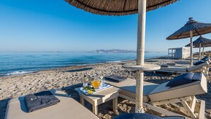 A beachside lounge with sun loungers, a table with a drink and snacks, and a view of the ocean.