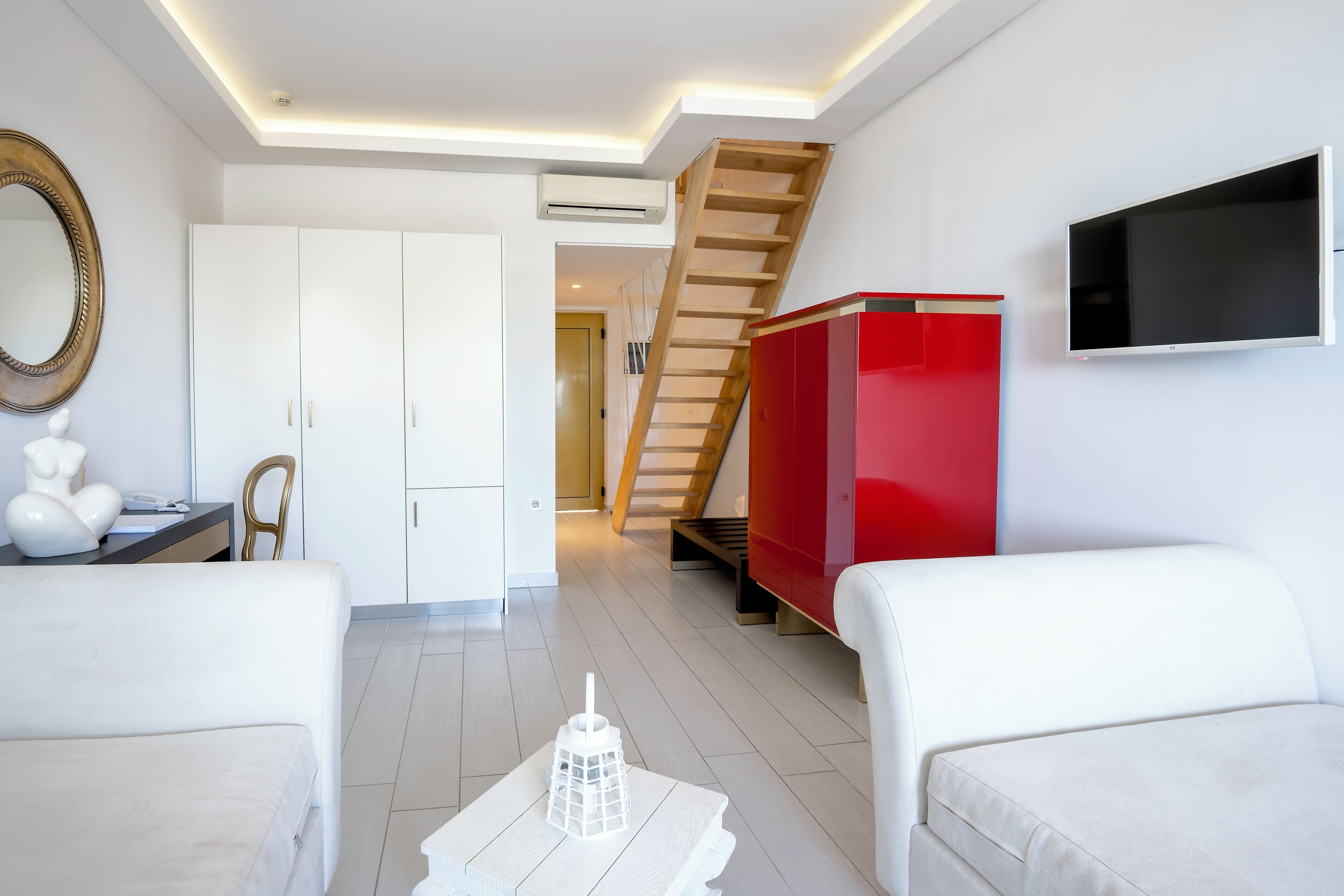 Una sala de estar moderna con sofás blancos, un aparador rojo, un televisor montado en la pared y una escalera que conduce a otra habitación.