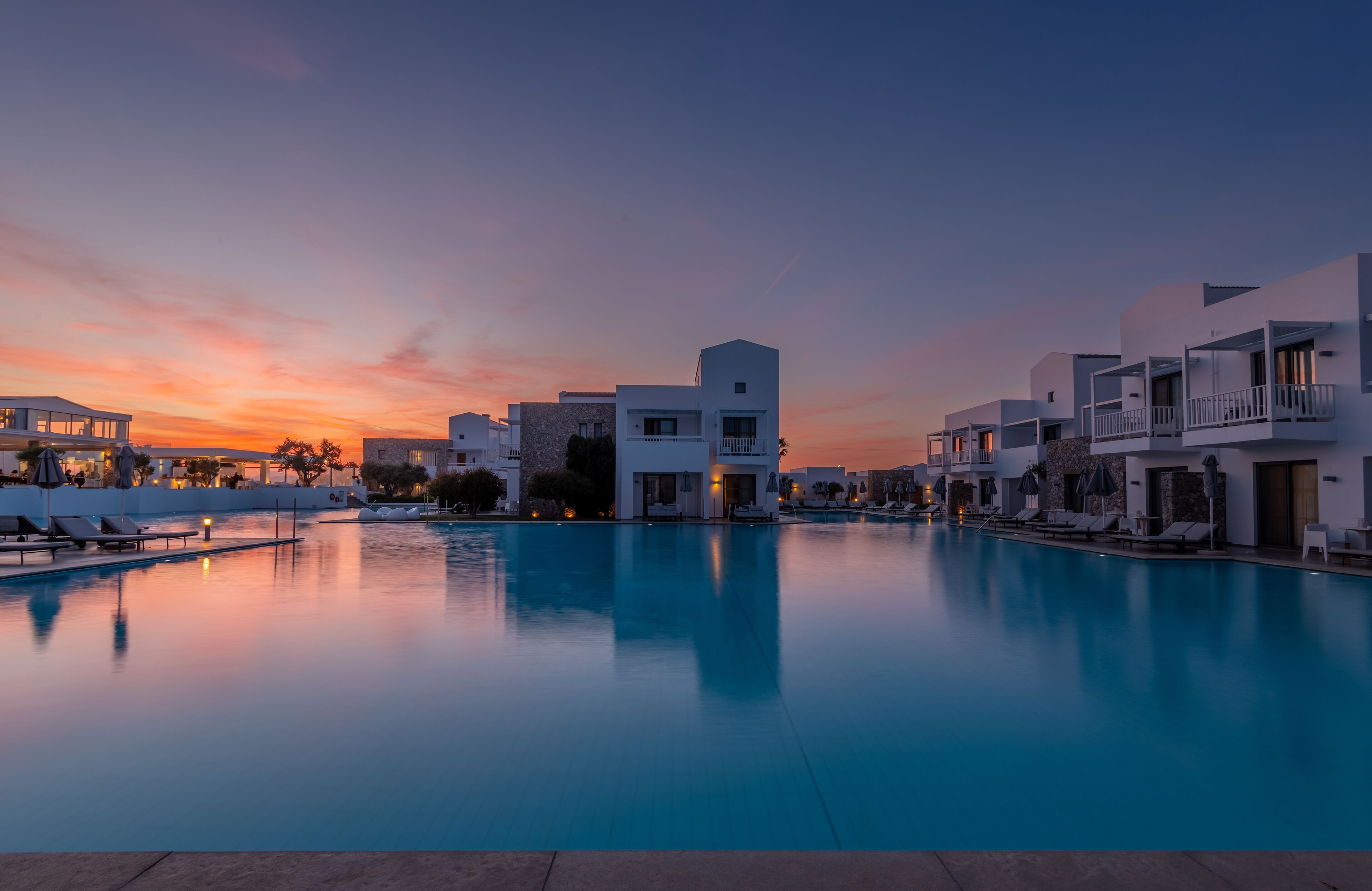 A modern hotel with a pool, white buildings, and a sunset sky.