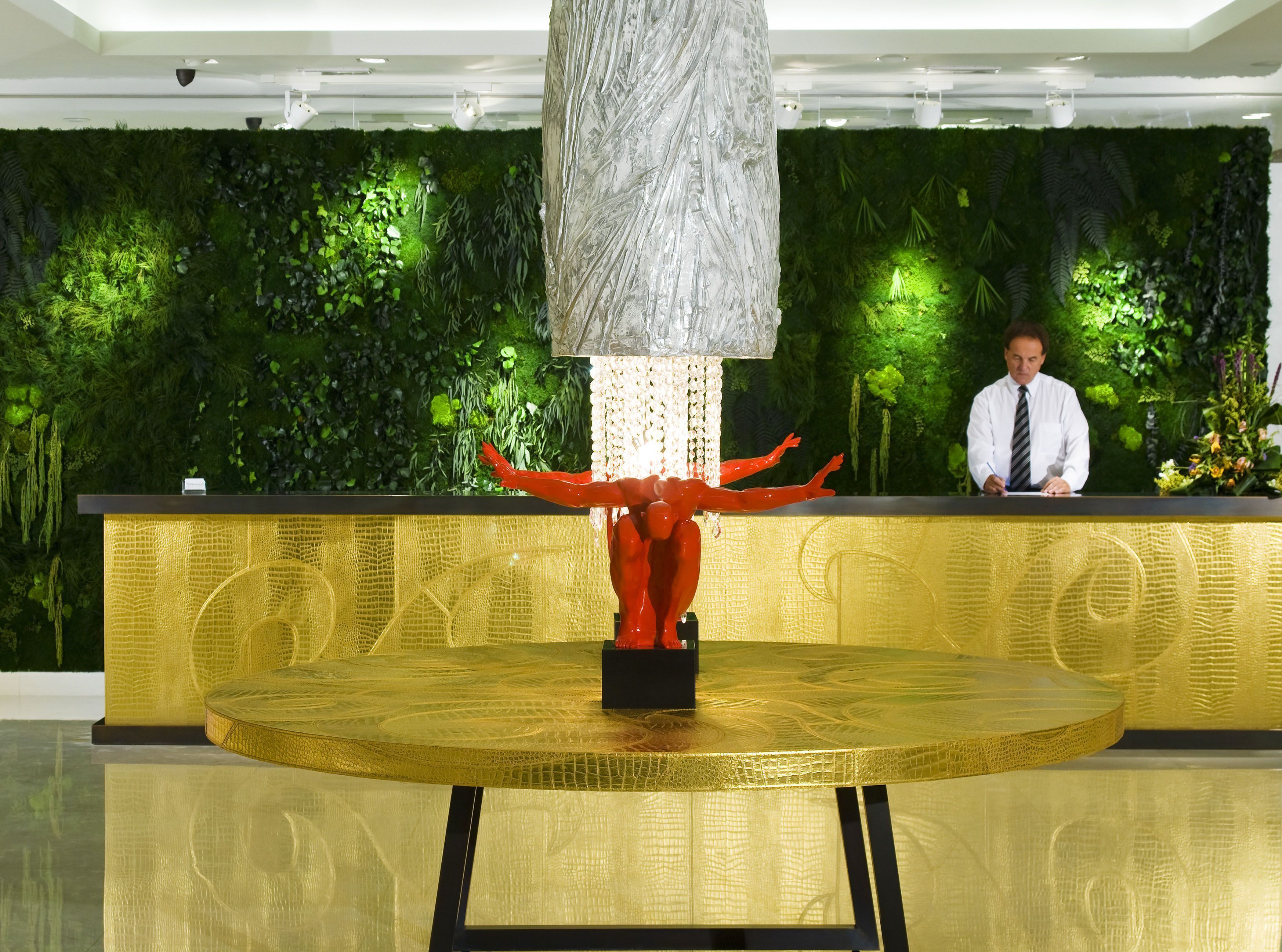 A reception desk with a golden textured surface, a large decorative sculpture in the center, and a green wall with plants in the background.