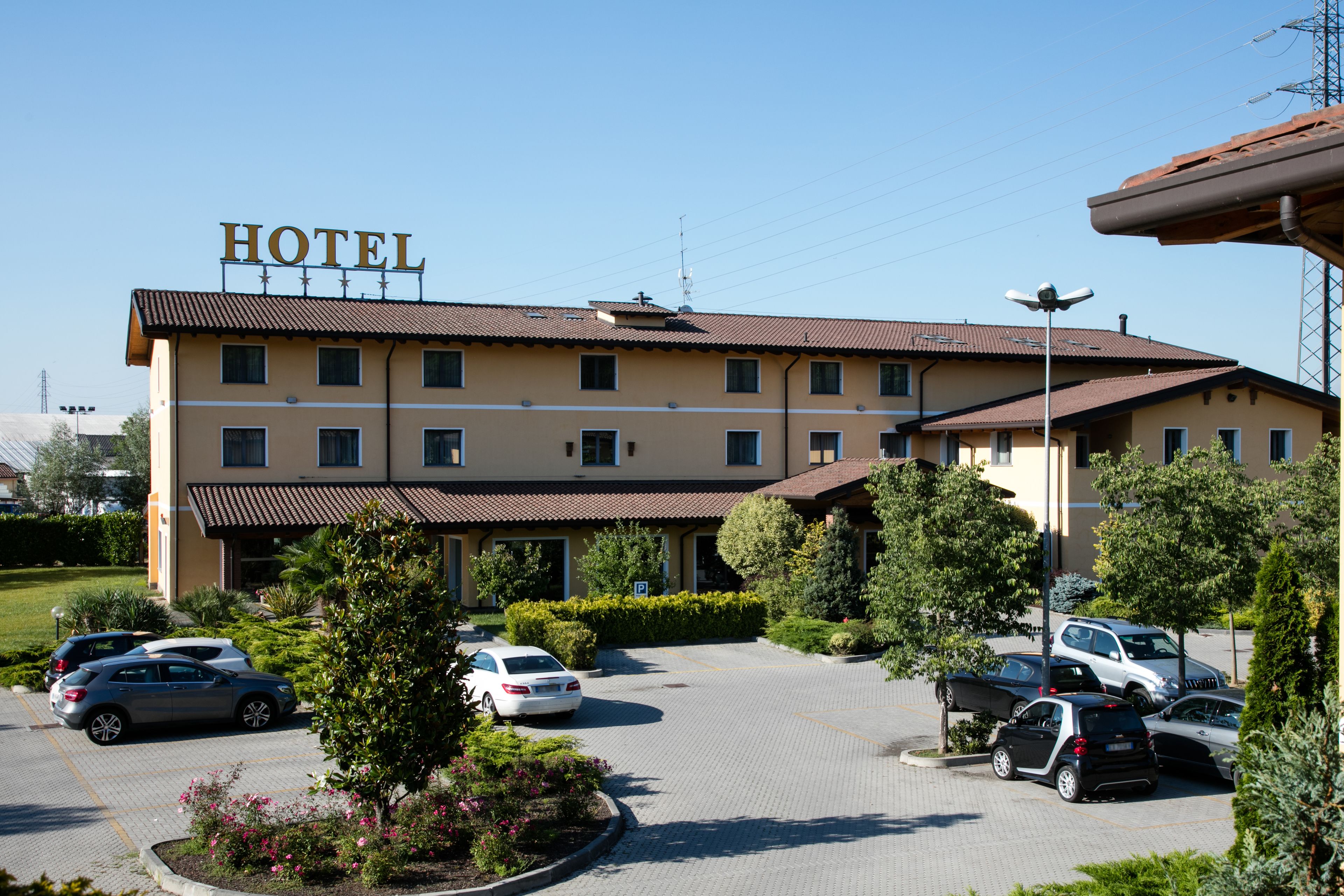 Ein Hotel mit einem Schild mit der Aufschrift 'HOTEL' auf dem Dach, einem Parkplatz mit mehreren Autos und einem begrünten Bereich mit Sträuchern und Blumen.