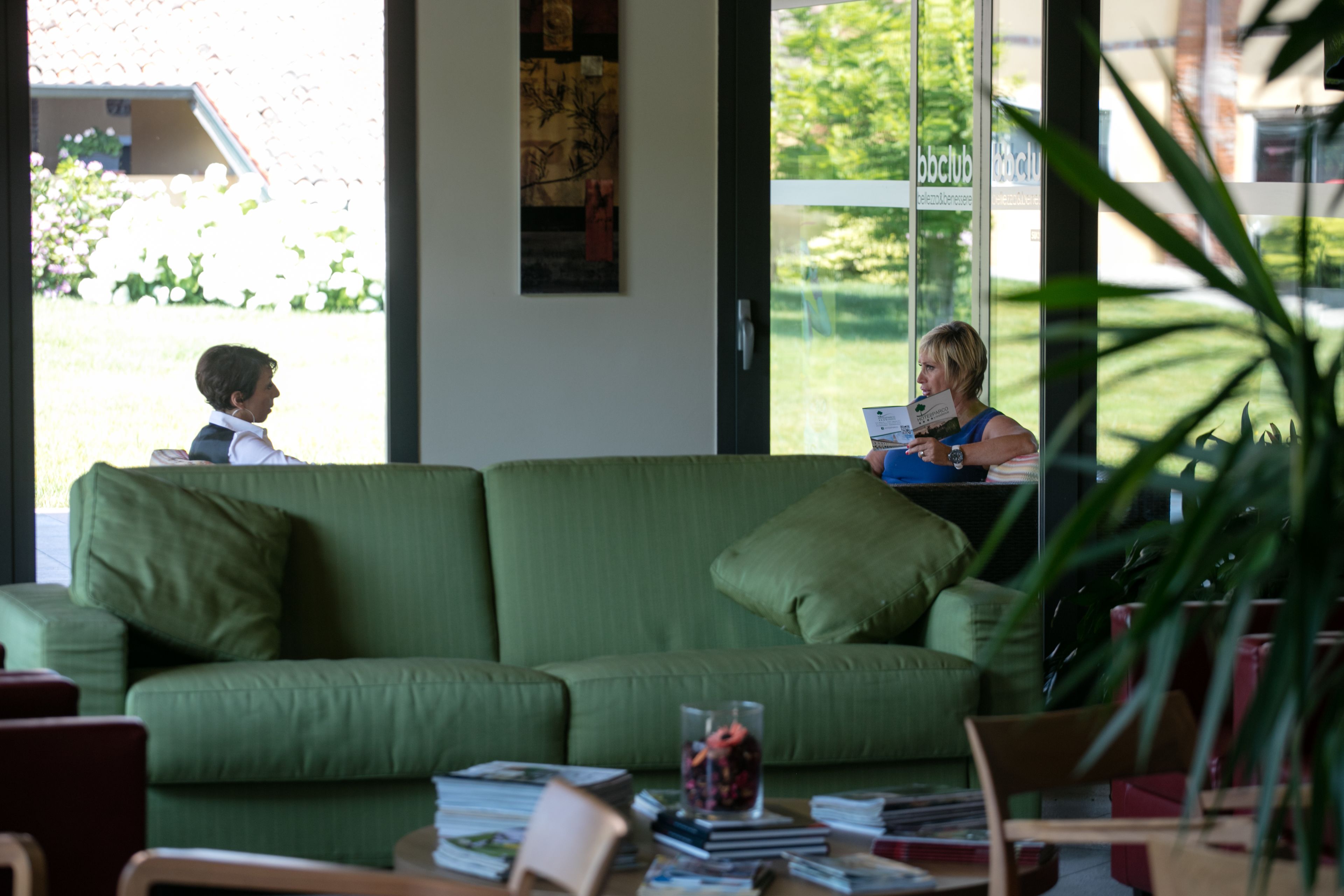 Een groene bank met een glazen vaas en tijdschriften, een houten tafel met boeken, en een raam met uitzicht op groen en een gebouw.