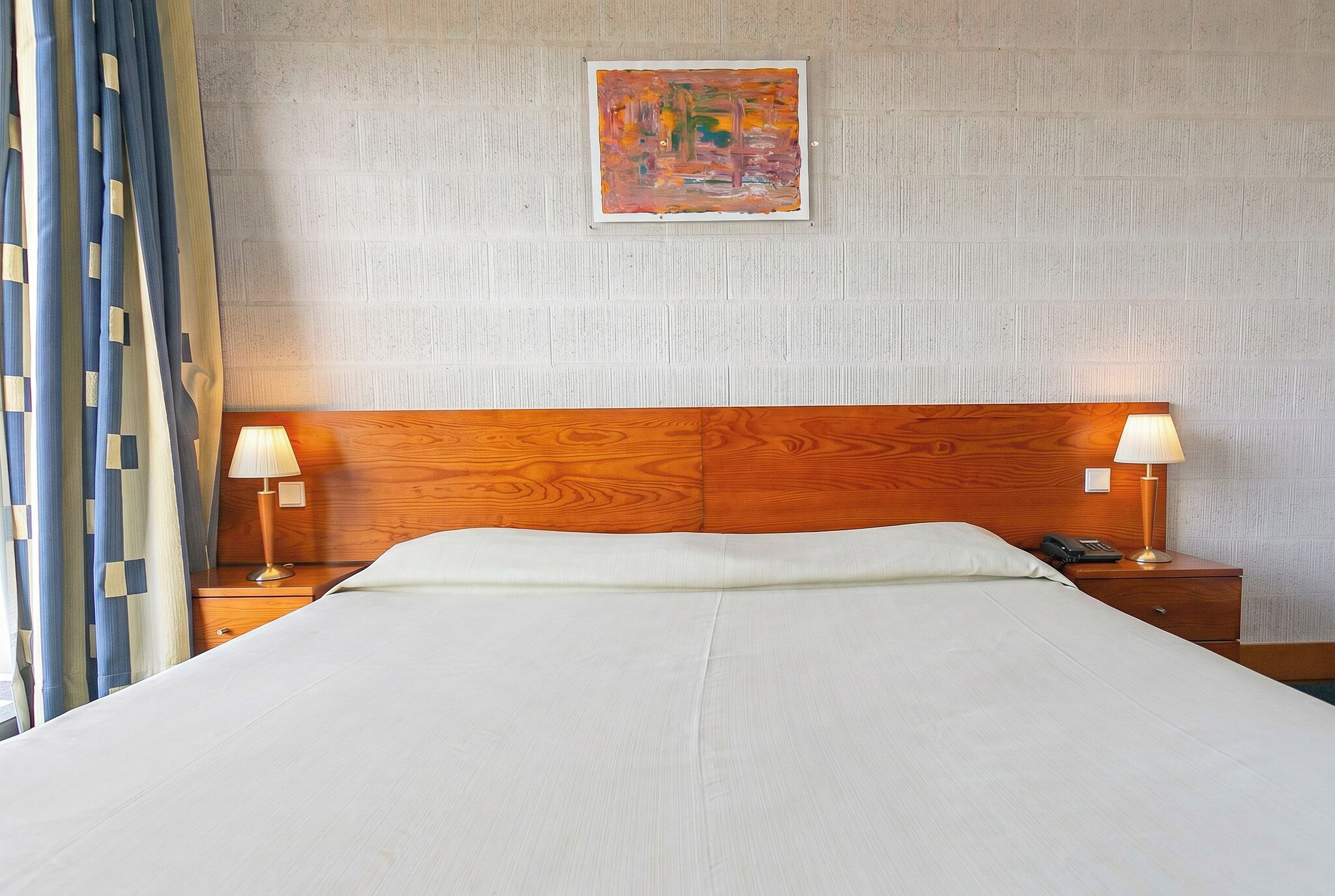 A hotel room with a wooden headboard, two bedside tables with lamps, a bed with white linens, a painting on the wall, and a curtain on the left side.