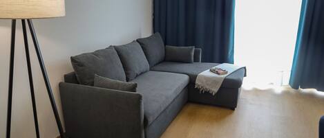 Un sofá seccional gris con un plaid, una lámpara de pie y una ventana con cortinas azules.