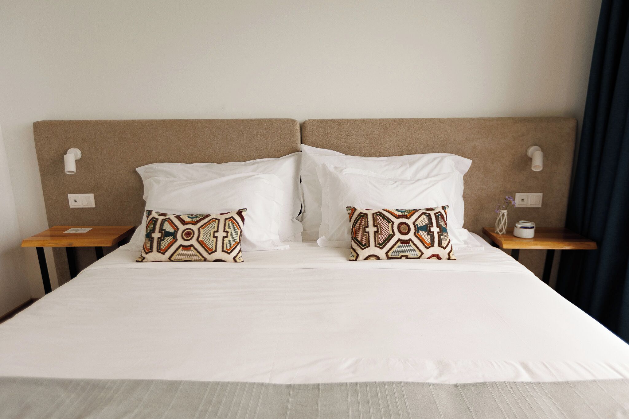 A hotel room with a large bed, two patterned pillows, a wooden nightstand, a small table, and a wall-mounted light.