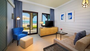 A hotel room with a sofa, a chair, a TV on a wooden cabinet, and a view of the beach. - Rixos Al Mairid Ras Al Khaimah (Ras Al Khaimah)