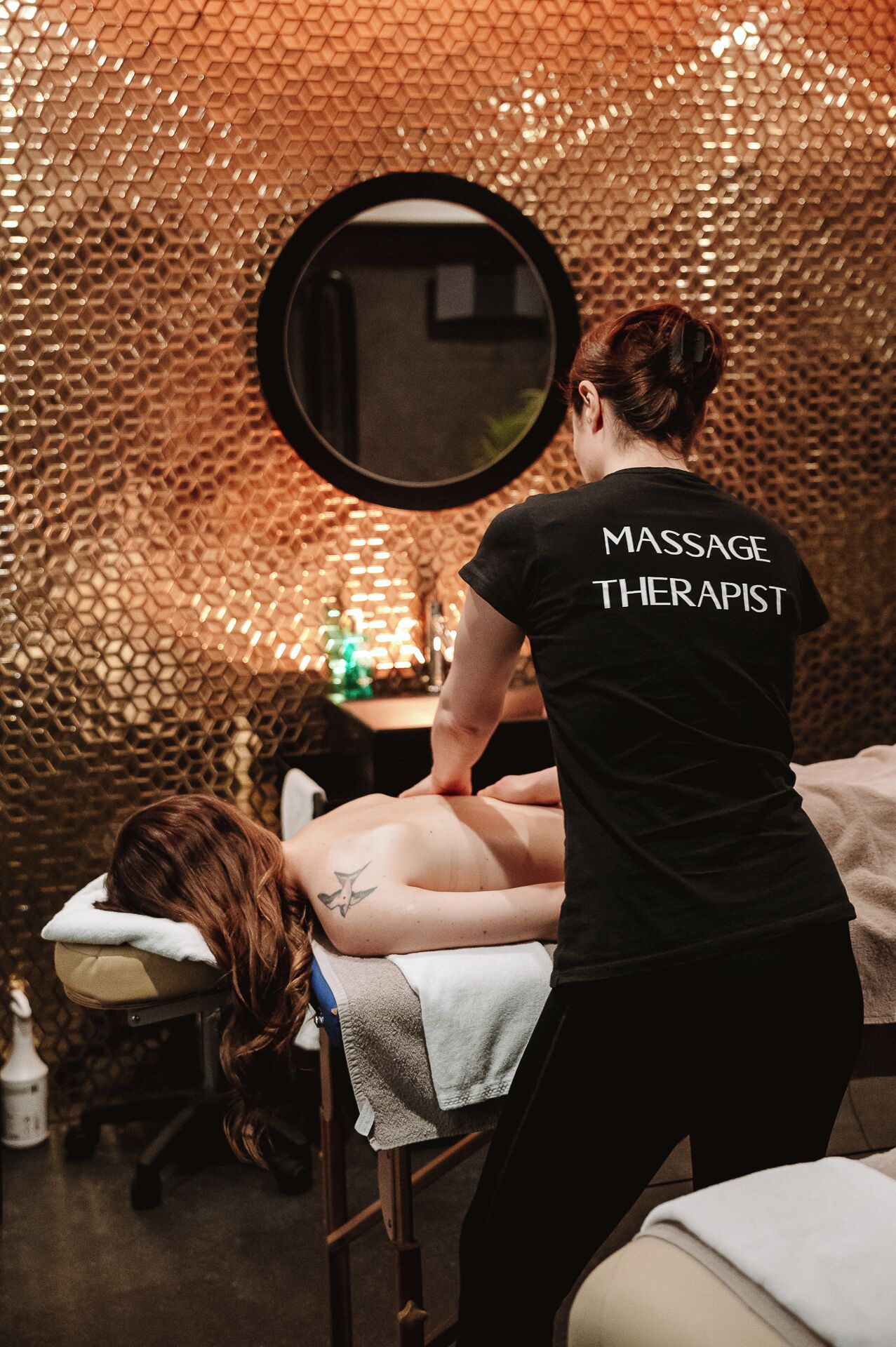 A massage therapist attending to a client in a room with a textured wall and a round mirror.