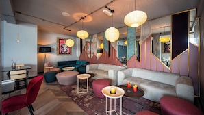 Un salon moderne avec un canapé d’angle, des fauteuils rouges et une table basse. On y trouve plusieurs luminaires suspendus et un grand miroir.