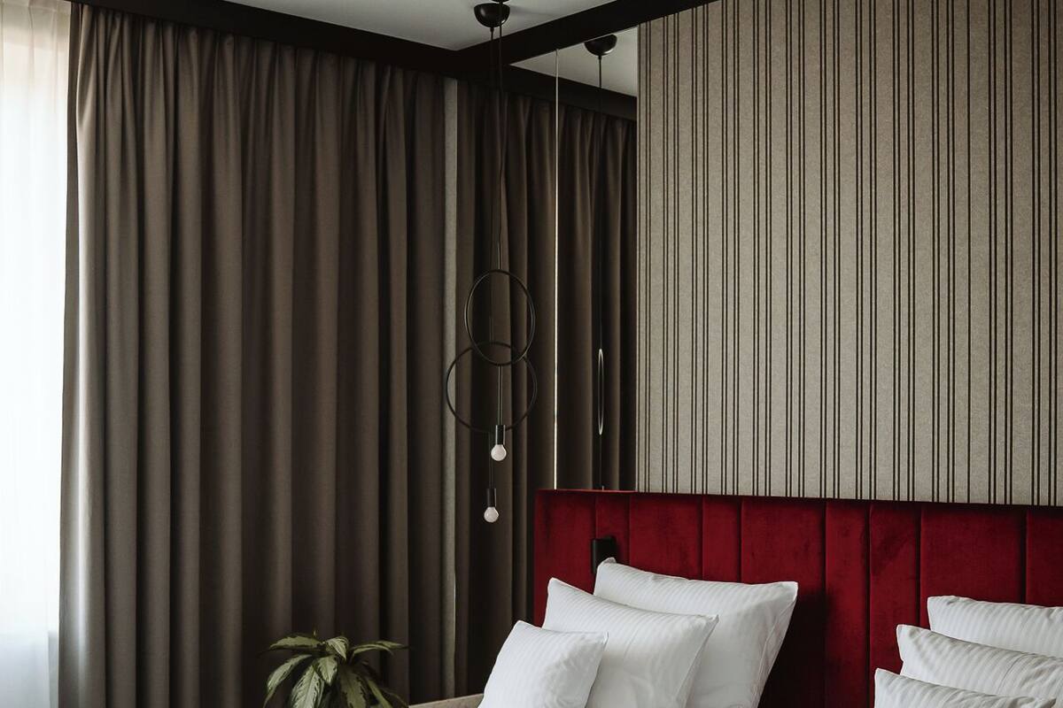 A hotel room with two beds, a red headboard, a large window with curtains, and a plant.