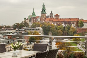 Un balcon doté de fauteuils et d’une table en osier, offrant une vue sur un paysage urbain avec des bâtiments historiques et un château.