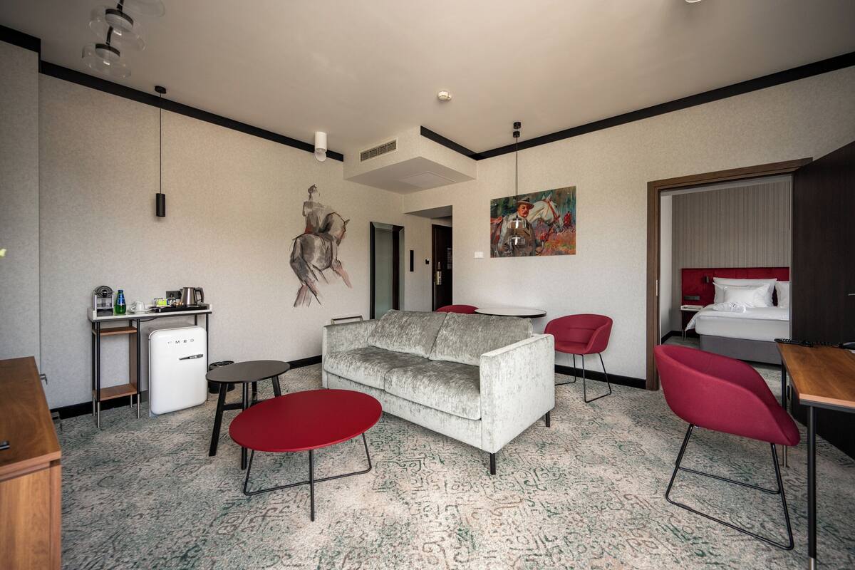 A hotel room with a sofa, two red chairs, a small table, a mini-fridge, a bed with a red headboard, and a painting on the wall.