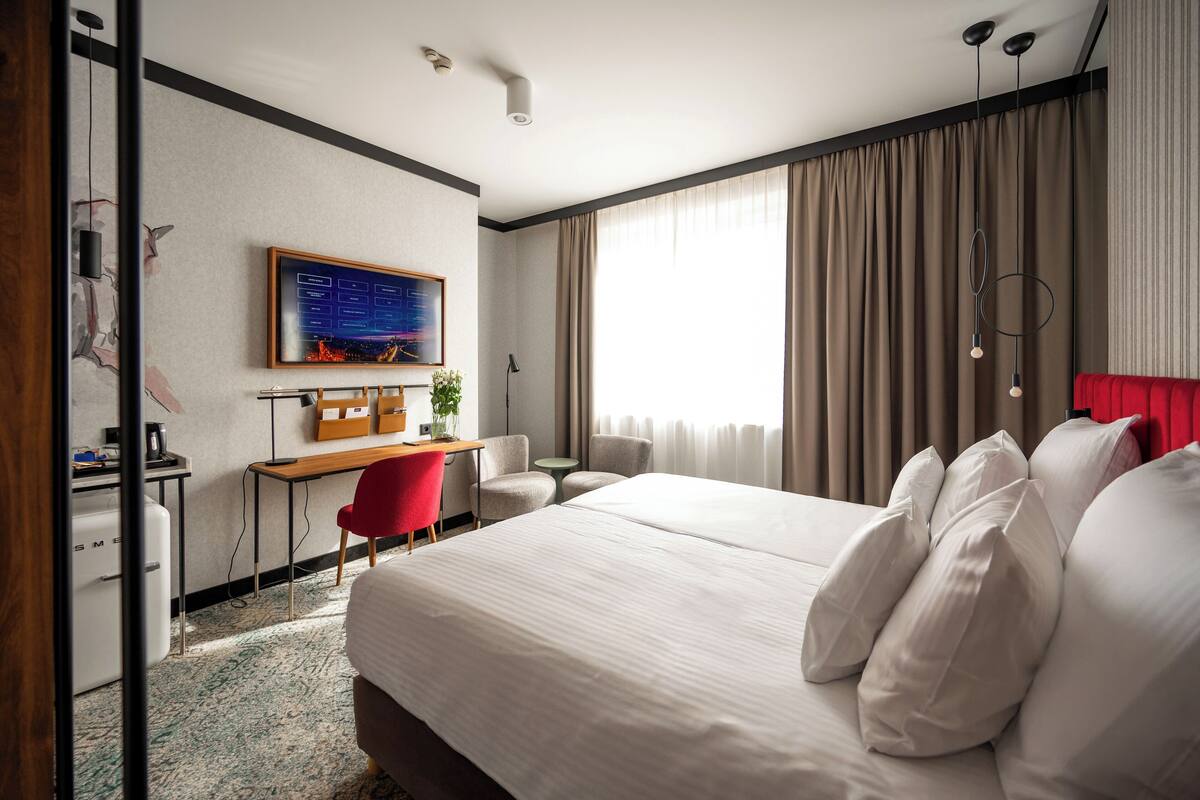 A hotel room with a large bed, a desk, a chair, a TV, and a window with curtains.