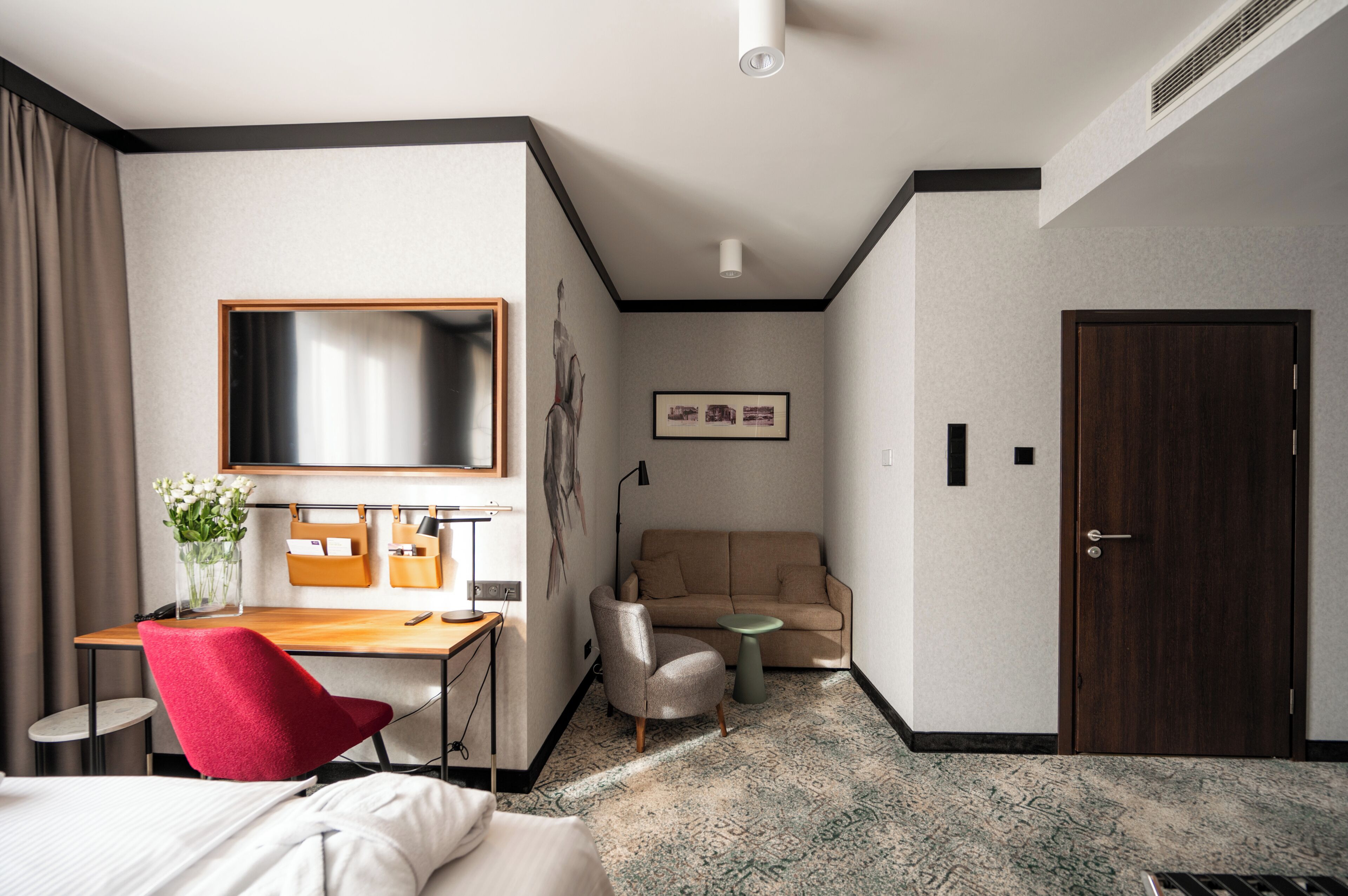 Habitación de hotel con cama, escritorio, silla, sofá, televisor, puerta y una obra de arte colgada en la pared.