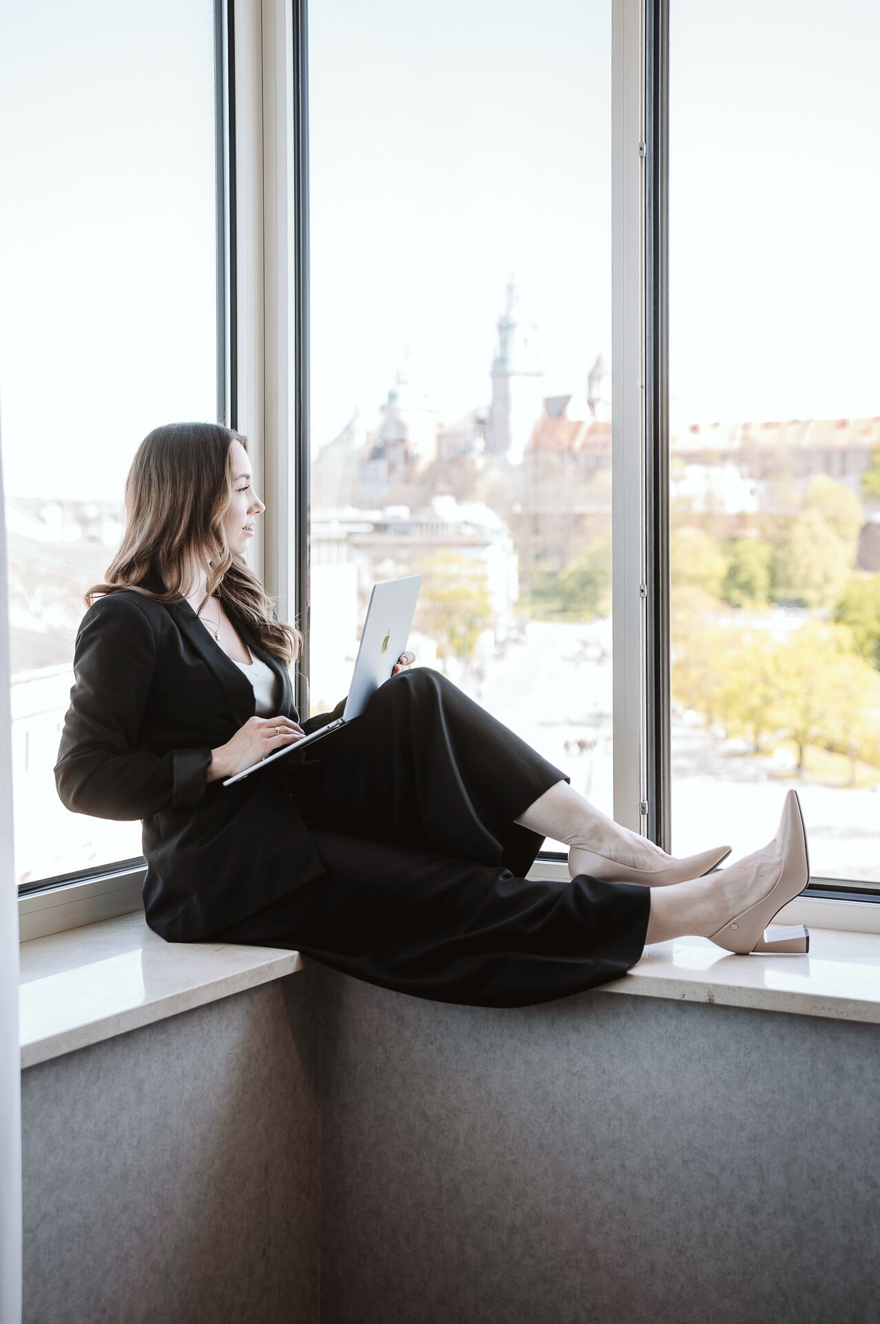 A person sitting by a window with a laptop, overlooking a cityscape with buildings and a river.