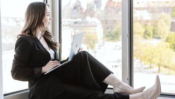 A person sitting by a window with a laptop, overlooking a cityscape with buildings and a river.