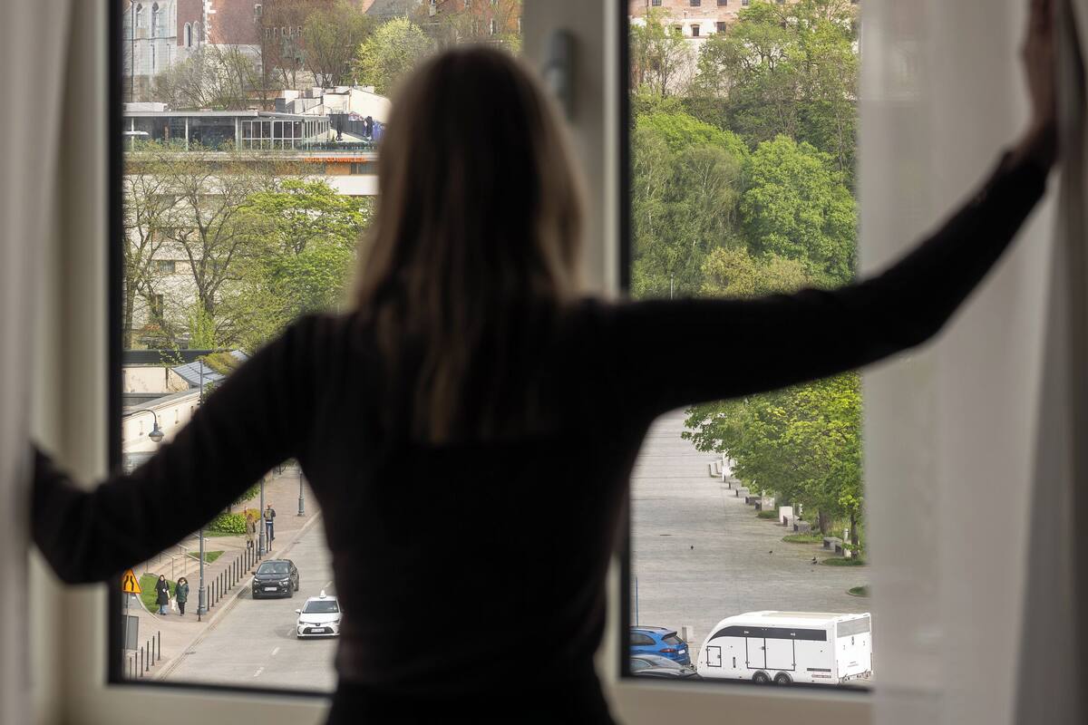 A person standing by a window with arms outstretched, overlooking a cityscape with historic buildings and a clear sky.