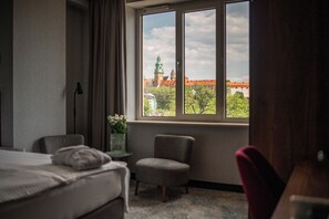 Une chambre d’hôtel avec un lit, une chaise, une petite table ornée d’un vase de fleurs et une fenêtre donnant sur un bâtiment doté d’une tour horloge.