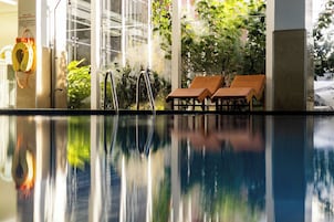 A pool with clear water reflecting the surroundings, a glass door entrance, and two wooden lounge chairs.