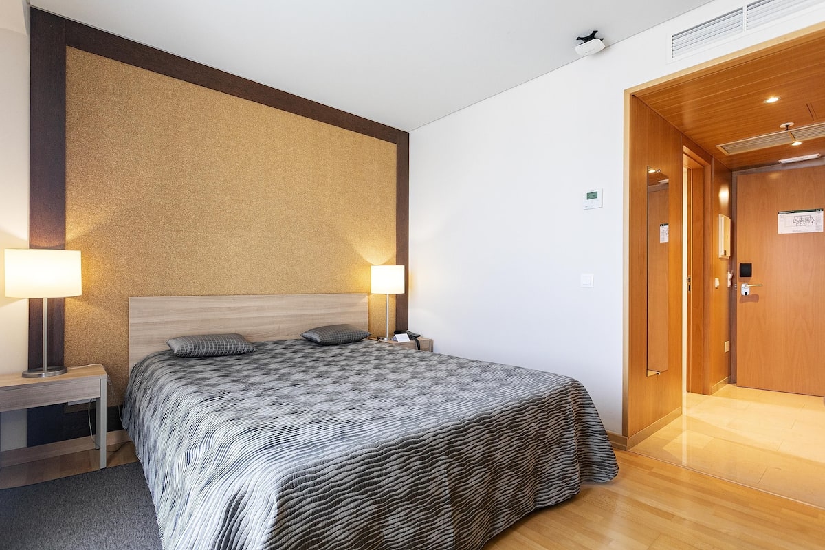 A hotel room with a large bed, two bedside lamps, a wooden headboard, and a wooden door.