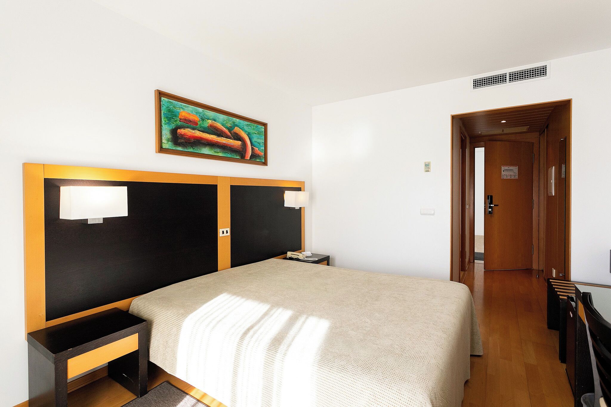 Una habitación de hotel con una cama, mesitas de noche, un cuadro y una puerta que da a otra habitación.