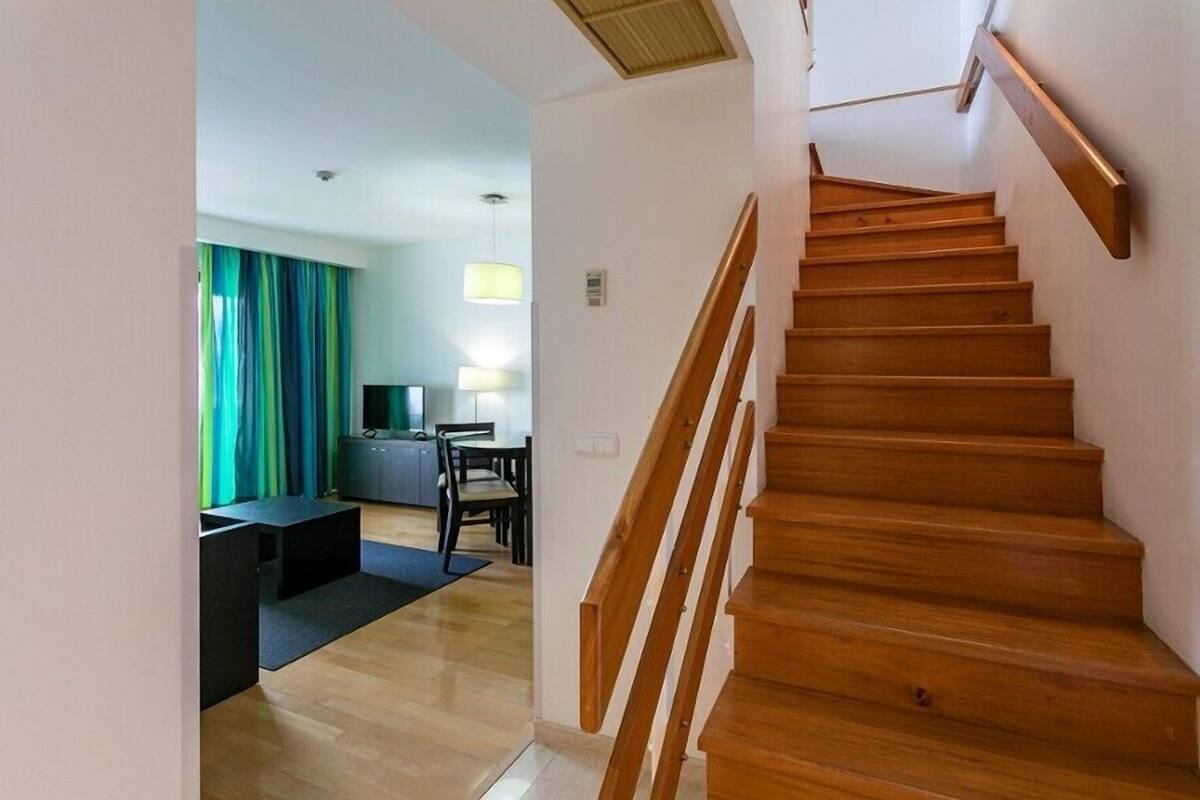 A staircase leading to an upper floor, a dining area with a table and chairs, and a view of a room with a bed and curtains.