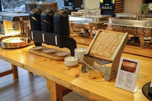 Montaje tipo bufé con varias estaciones de comida, incluyendo una plancha, un dispensador de miel y un cartel con los artículos del menú.