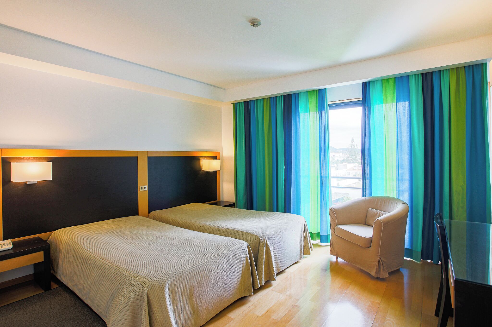 Ein Hotelzimmer mit zwei Betten, einem Sessel, einem Nachttisch und einem Fenster mit Vorhängen.