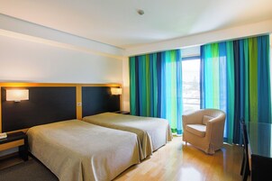 Una habitación de hotel con dos camas, una silla, una mesita de noche y un ventanal con cortinas.