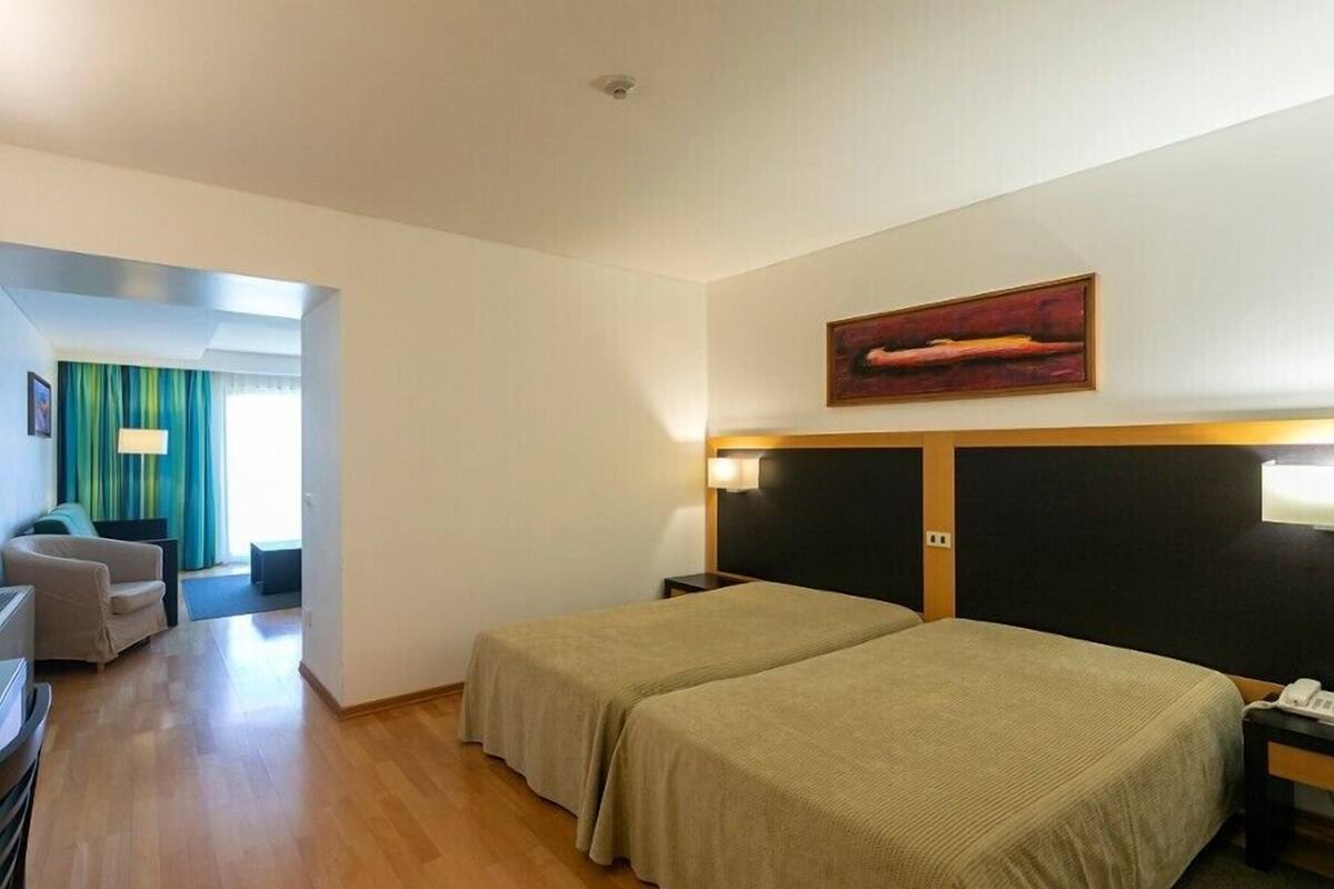 A hotel room with two beds, a desk, a chair, a painting, and a view of the hallway.