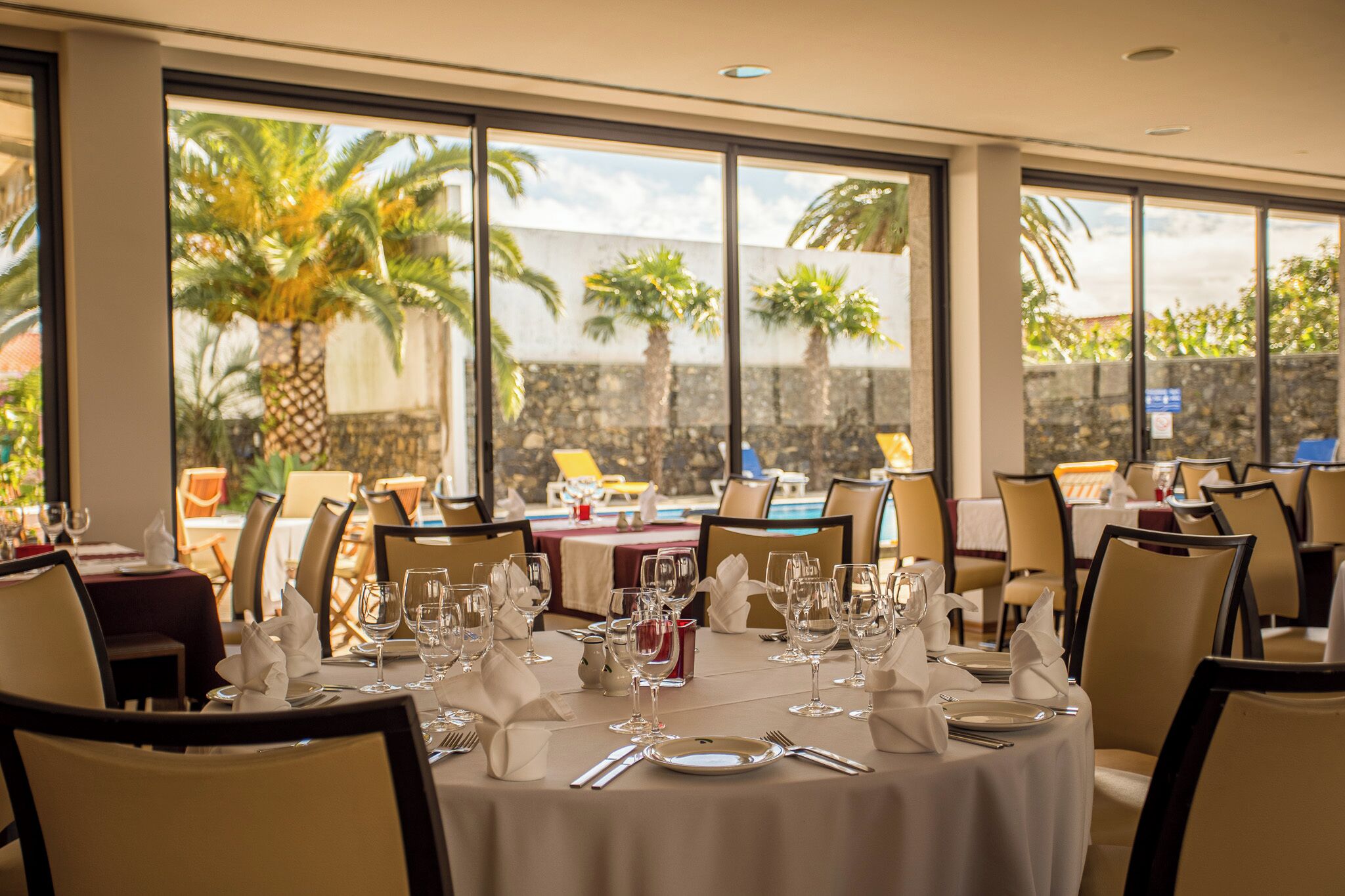 A restaurant with round tables set for dining, large windows with a view of palm trees, and a stone wall in the background.