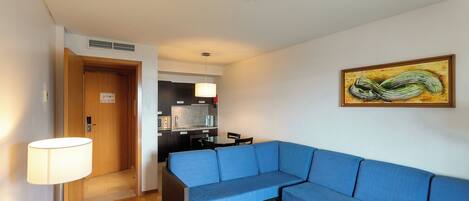 A modern hotel room with a blue sofa, a dining area, and a kitchen.