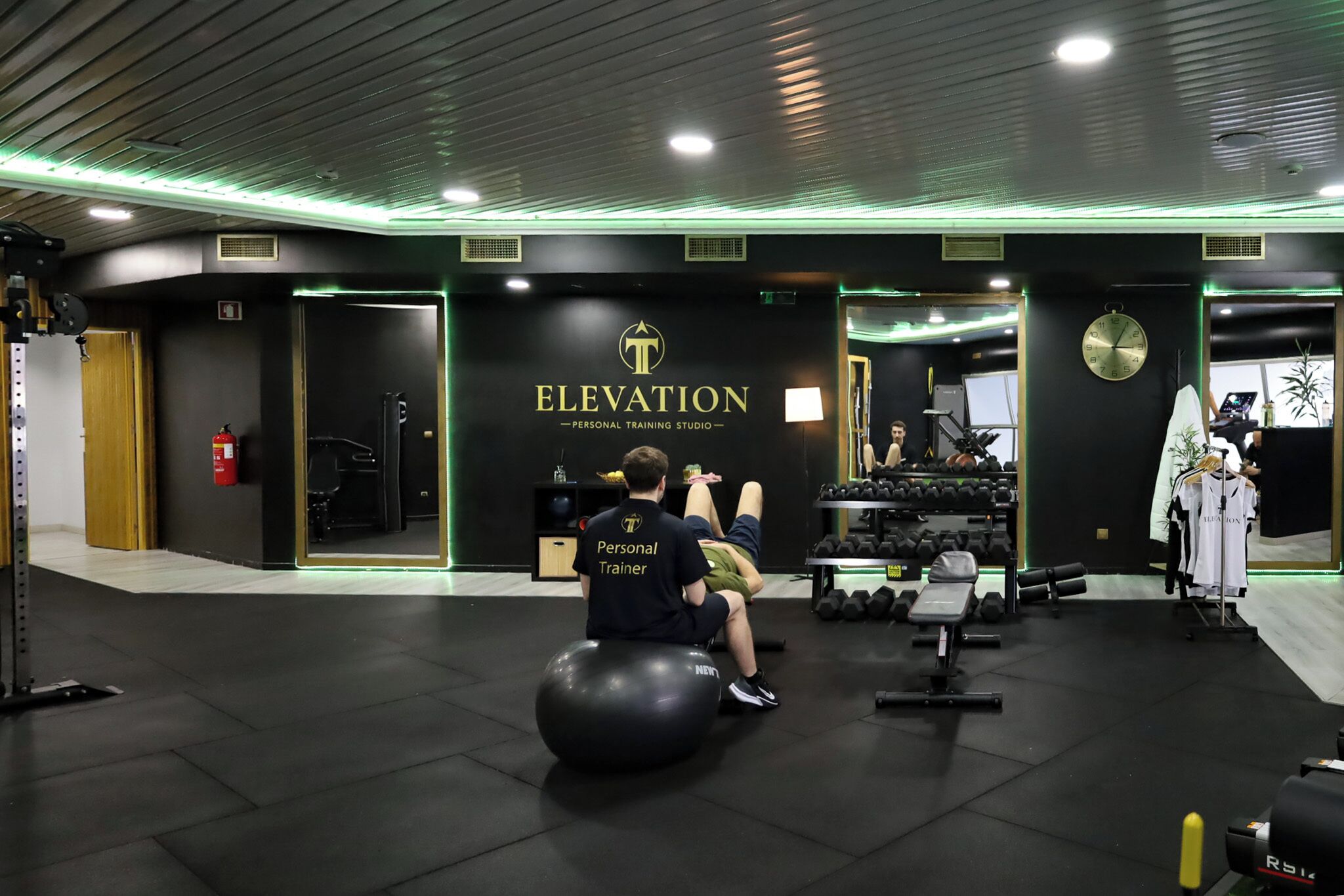 Ein Fitnessstudio mit Schwarz- und Goldtönen, mit einer Wanduhr, Kurzhanteln und einem sitzenden Trainer.