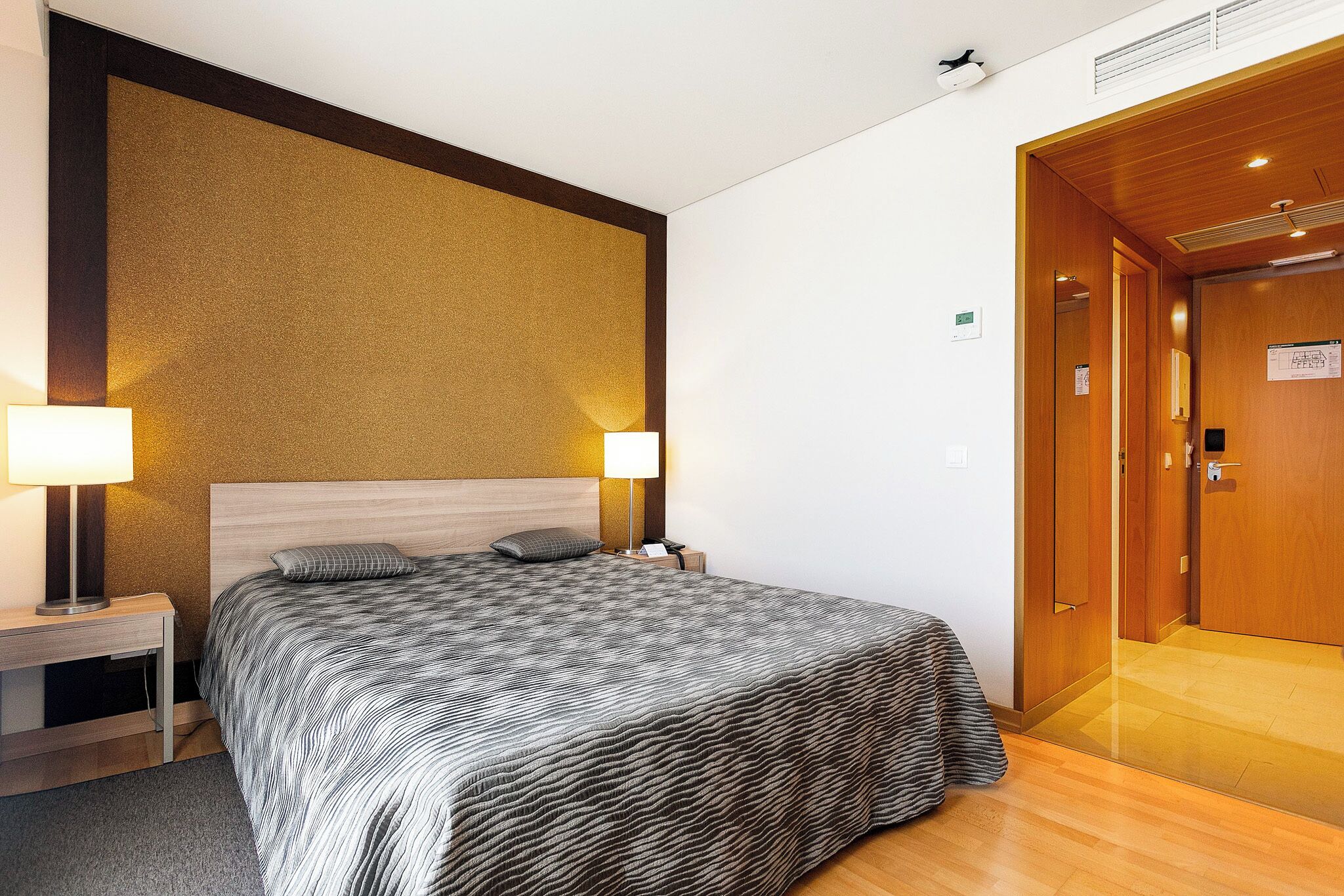 Een hotelkamer met een bed, nachtlampjes, een nachtkastje, een houten deur en een muur met een structuur.