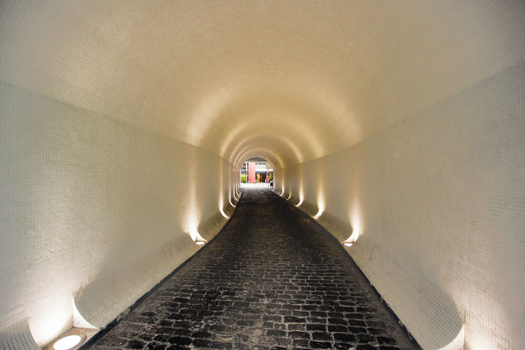 Un passage en forme de tunnel, avec un sol en briques et des murs blancs.