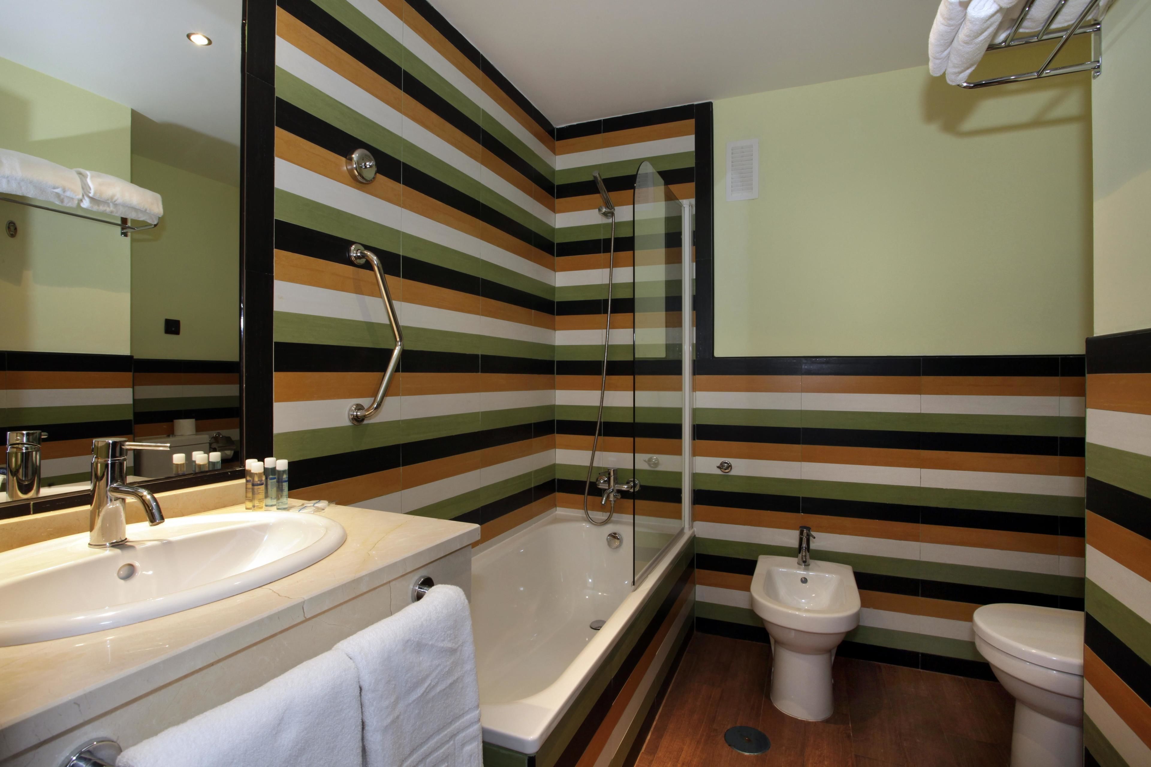 A bathroom with a striped tile wall, a bathtub, a sink, a bidet, and a mirror.