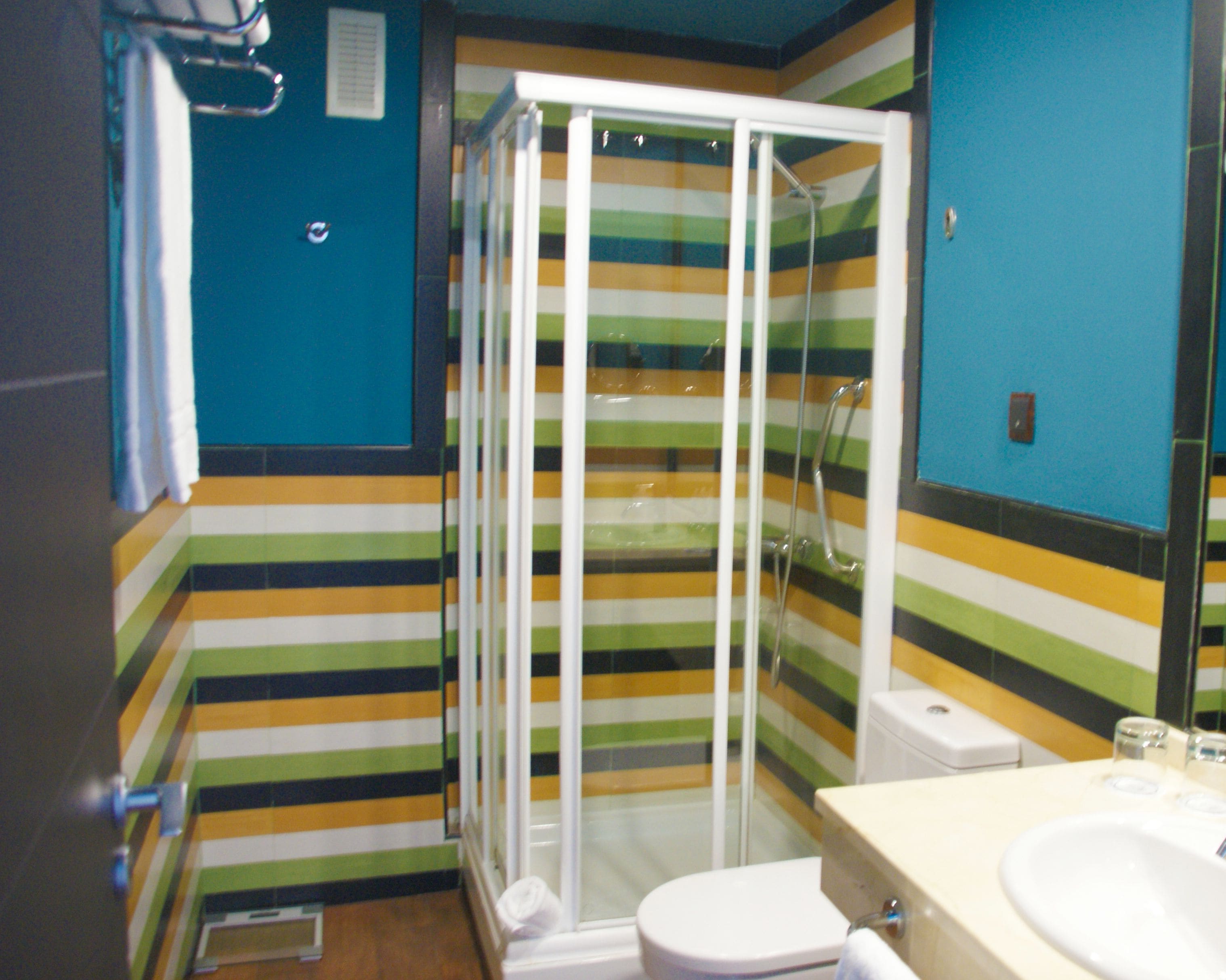 Un baño con piso a rayas, cabina de ducha de vidrio, inodoro blanco y lavamanos con espejo.