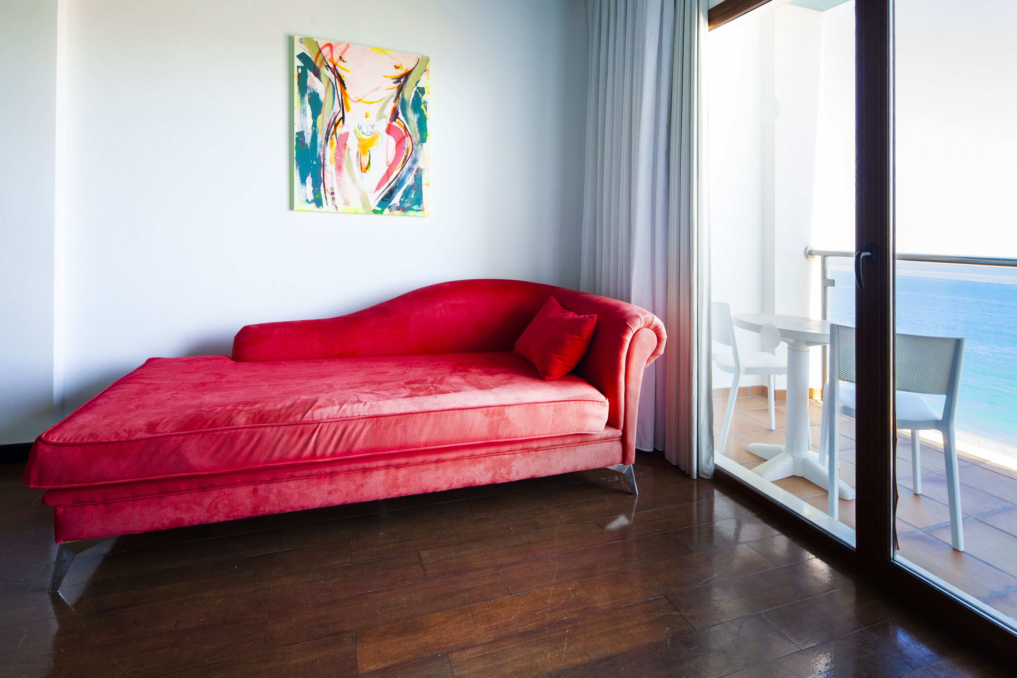 A red sofa in a room with a wooden floor, a white wall, and a colorful abstract painting.