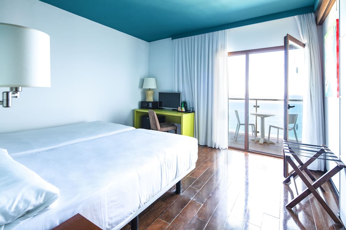 Une chambre d’hôtel avec un lit, un bureau doté d’une lampe, une chaise et une vue sur la mer, visible à travers une porte vitrée.