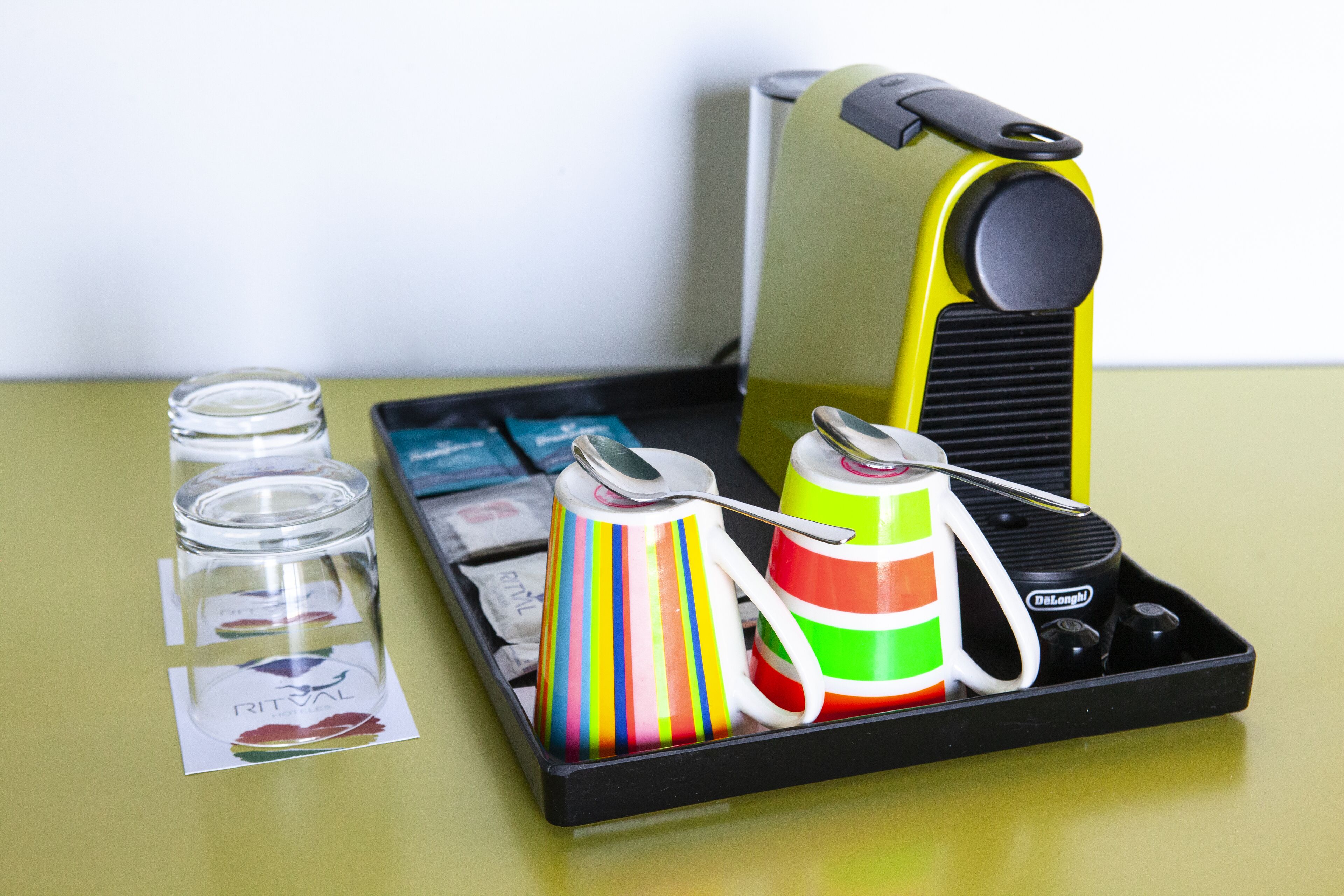 A tray with a coffee maker, colorful cups, a spoon, and a small container.