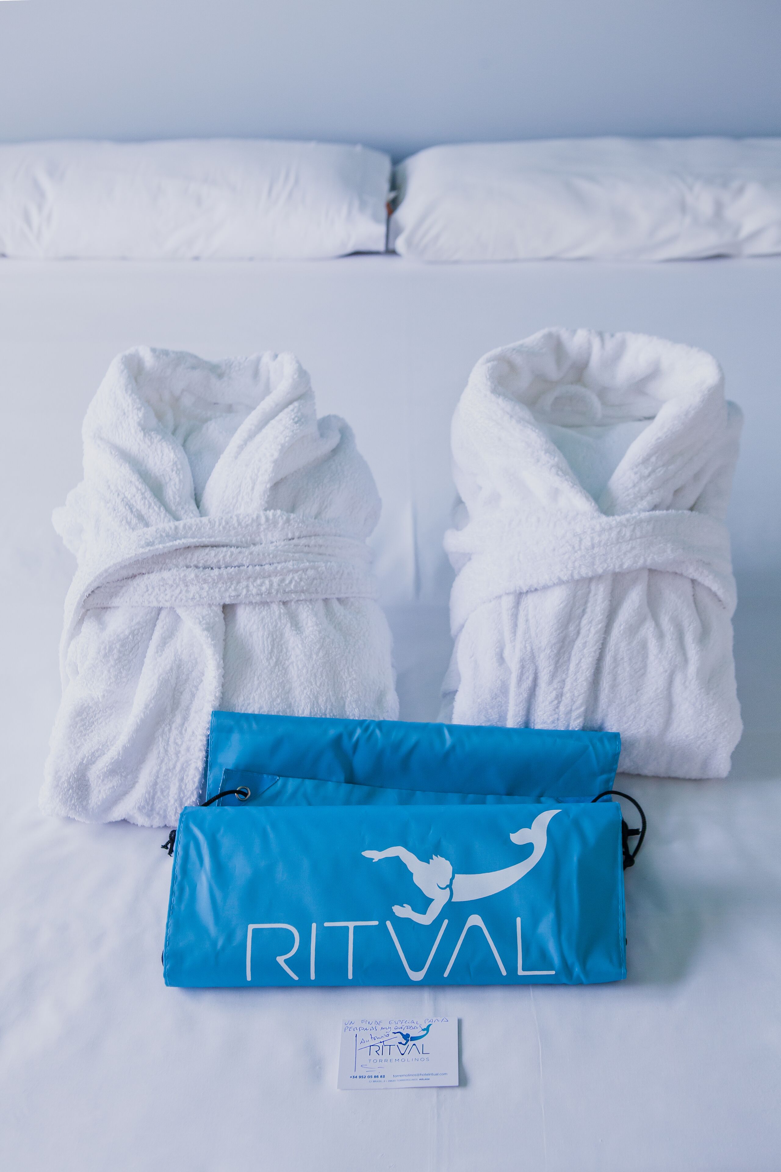 A neatly made bed with white pillows and two white bathrobes. A blue bag with the word "RITVAL" is placed on the bed.