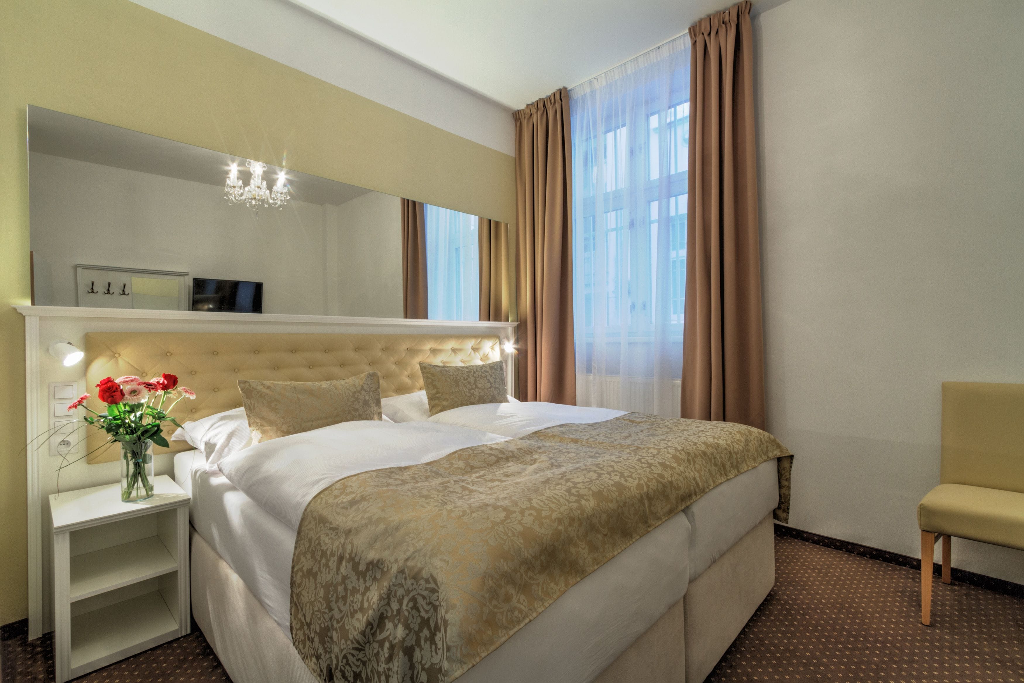 Habitación de hotel con una cama grande, una mesita de noche con un florero, una silla y un espejo sobre la cama.