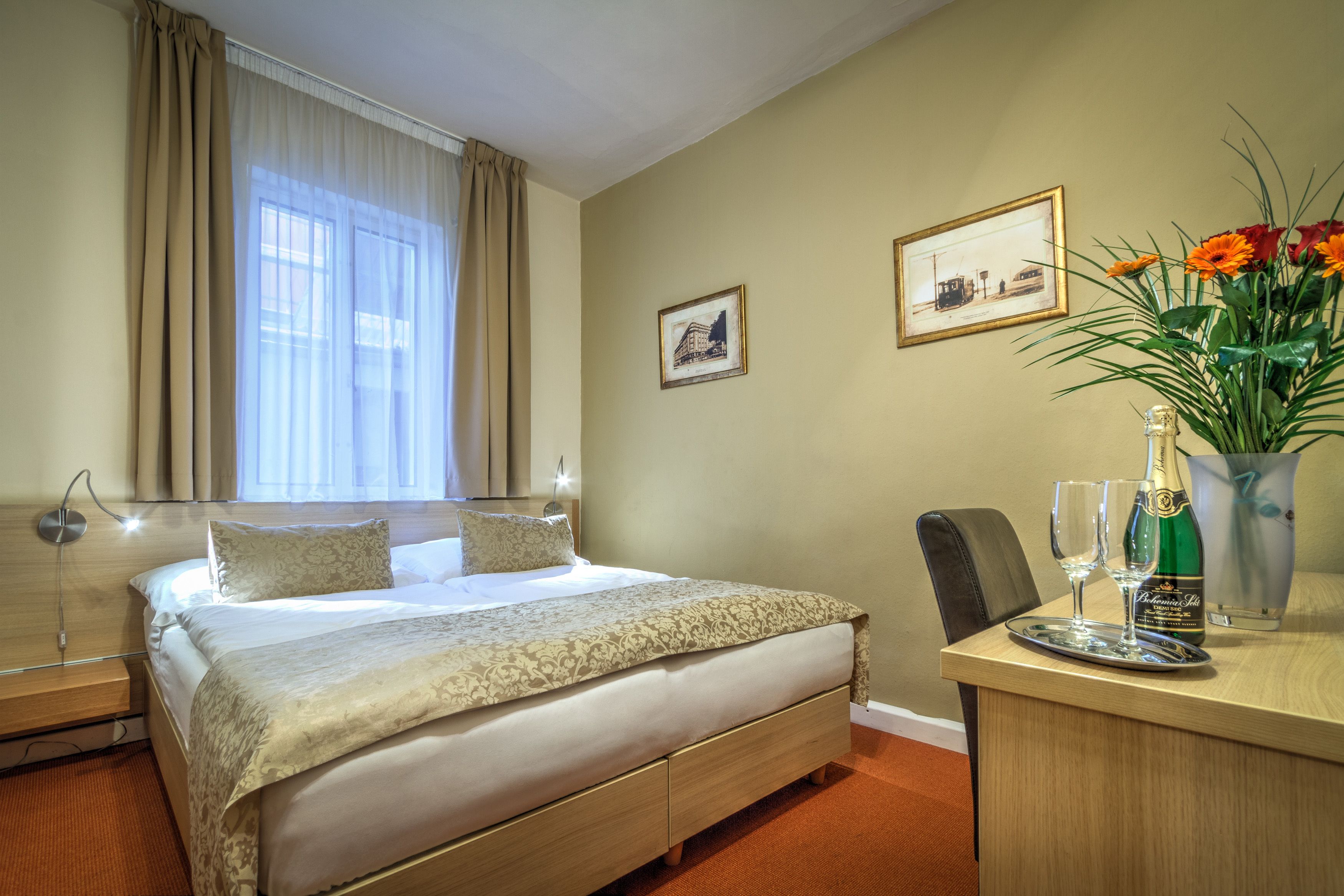 A hotel room with a bed, a desk with a bottle of champagne, a vase with flowers, and two framed pictures on the wall.