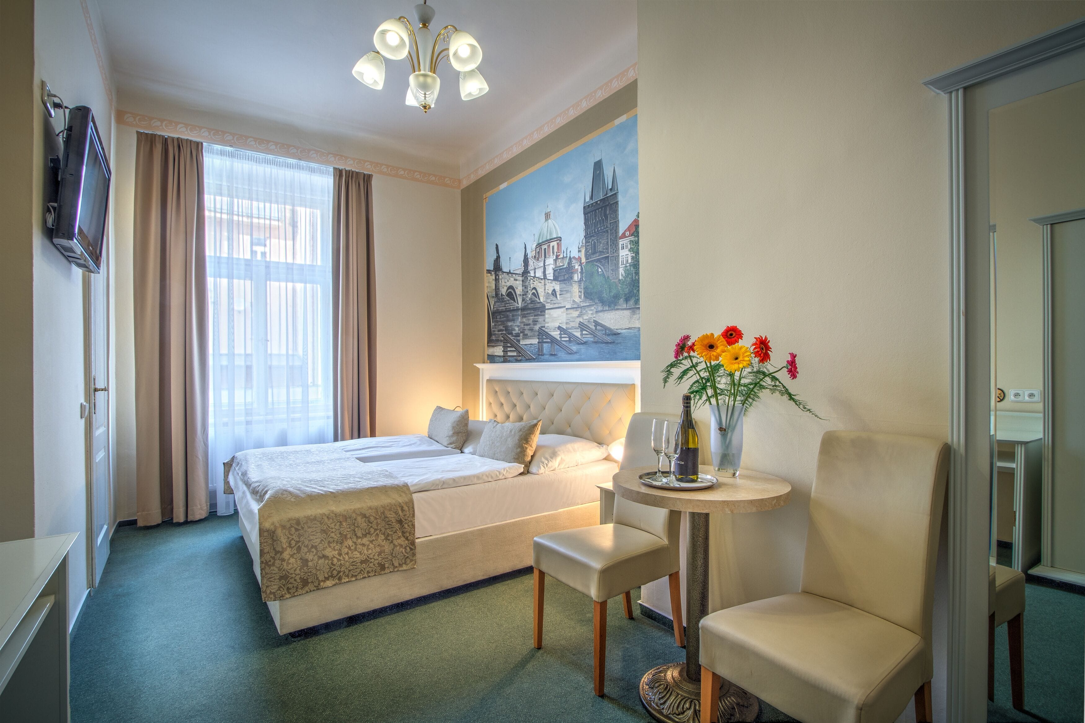 A hotel room with a bed, a chair, a small table with a bottle and glasses, a painting on the wall, and a television on the wall.