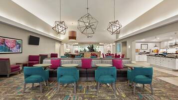 A modern hotel lobby with a seating area, a kitchen in the background, and geometric pendant lighting.