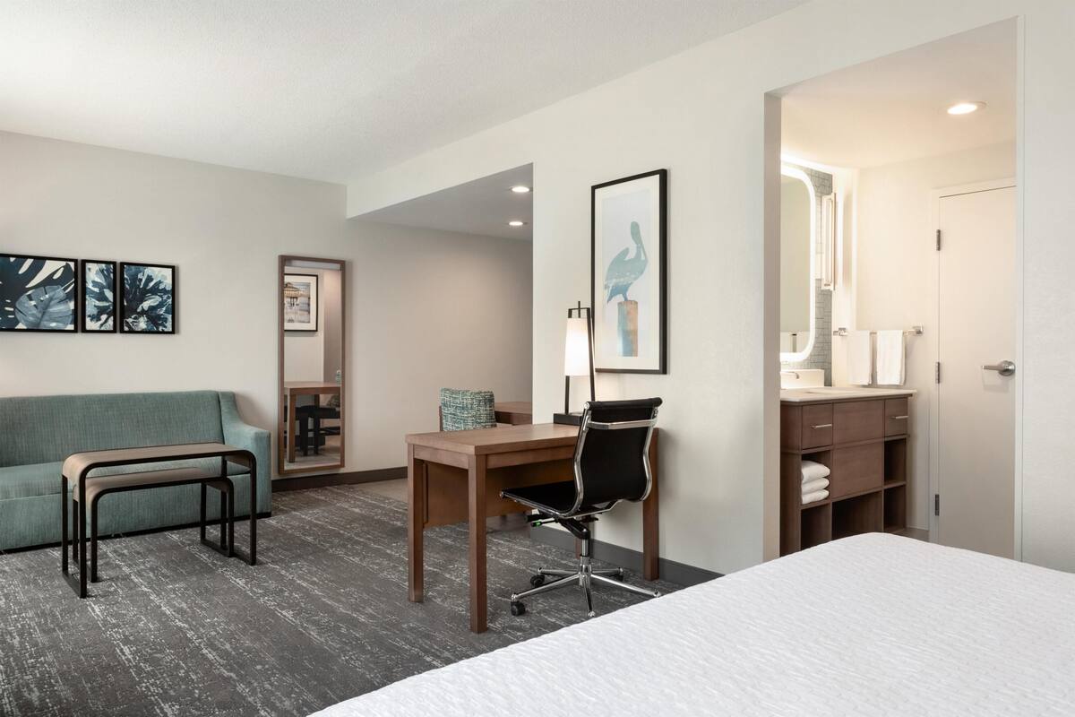 Una habitación de hotel con cama, escritorio, silla, sofá, espejo y obras de arte en las paredes.