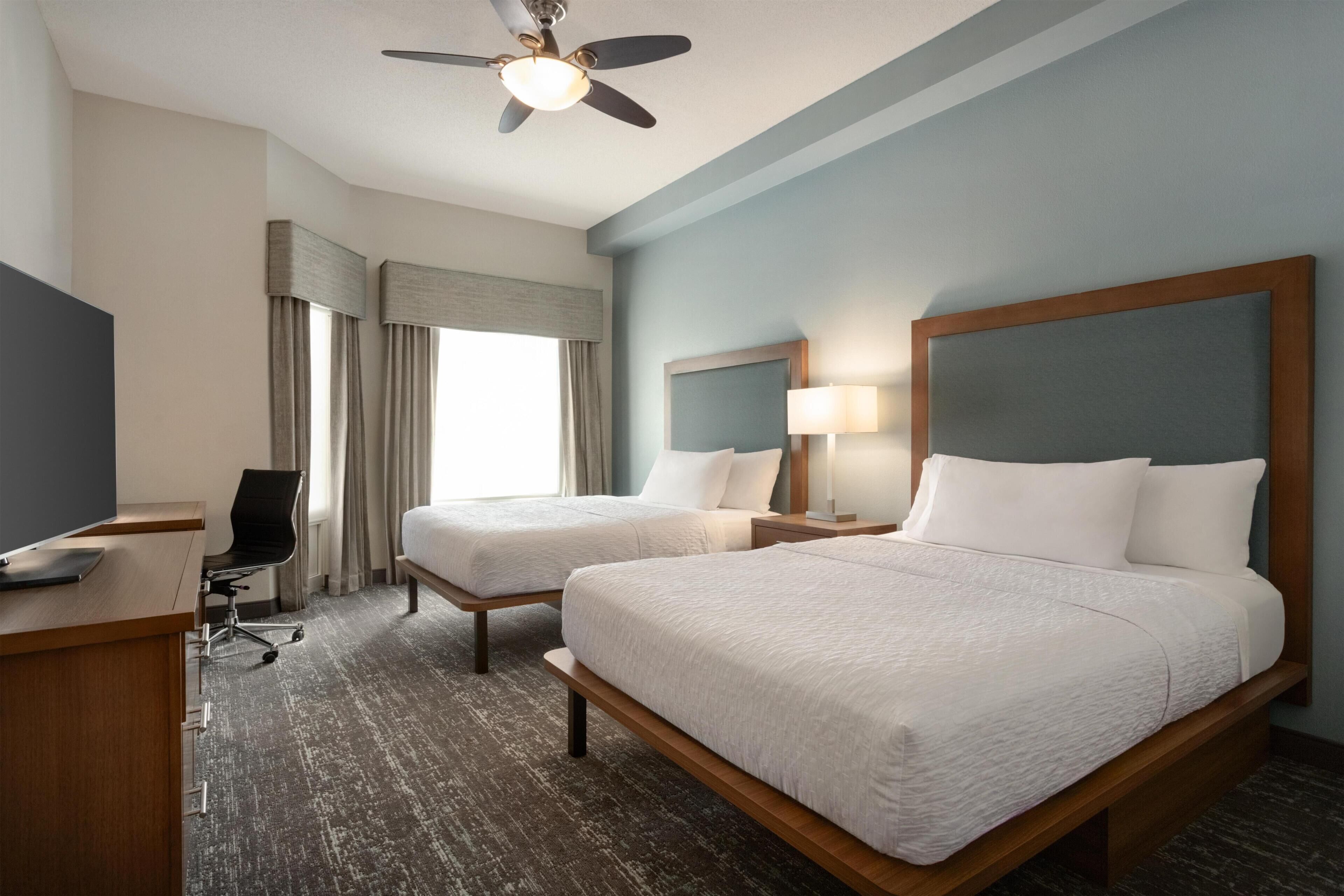 A hotel room with two beds, a desk, a chair, a ceiling fan, and a window with curtains.