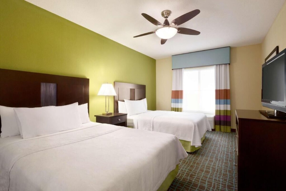 Una habitación de hotel con dos camas, ventilador de techo, televisor y ventana con cortinas.