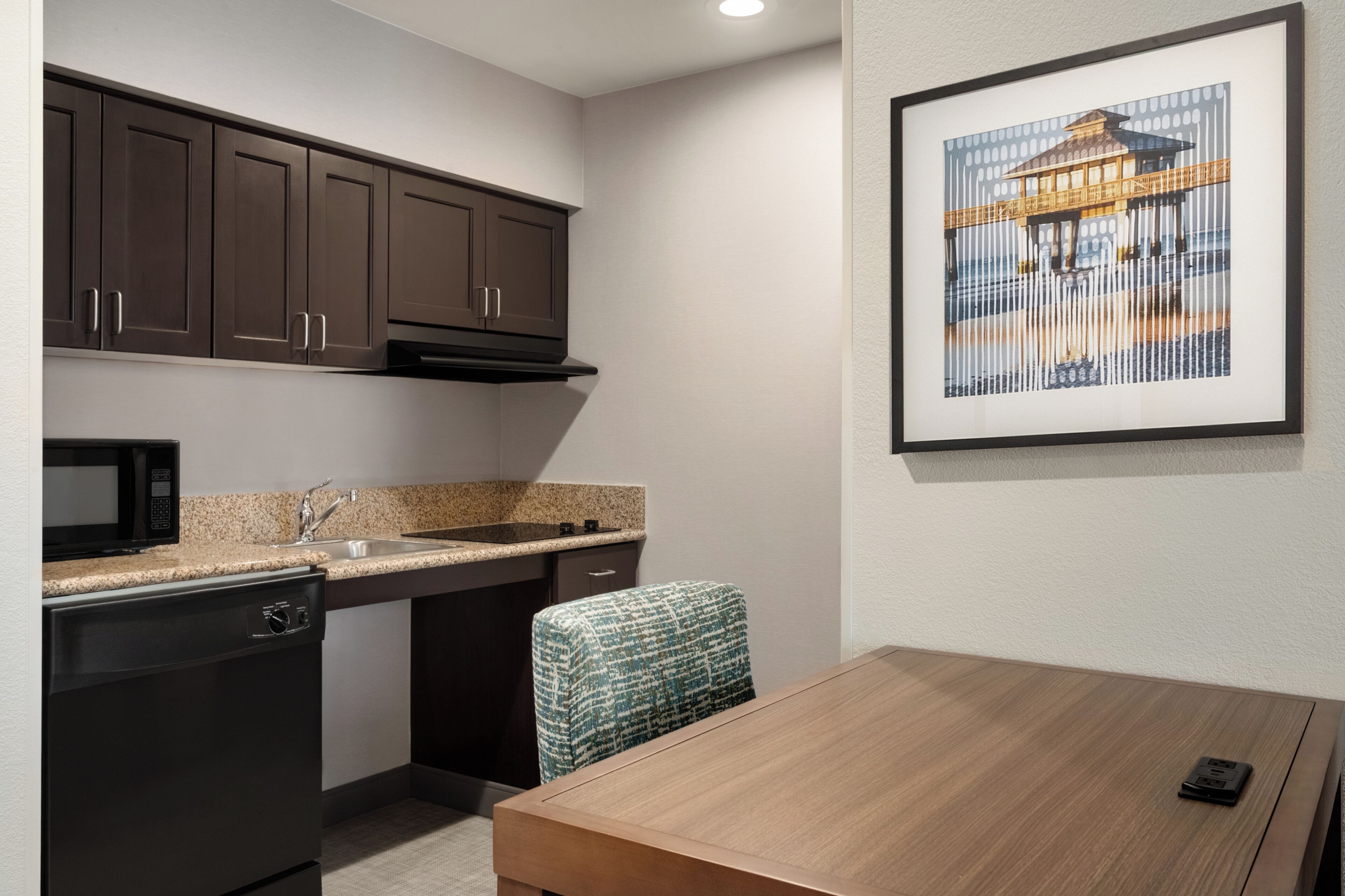 A compact kitchen with dark cabinets, a microwave, and a sink. There's a framed picture on the wall and a wooden table with a chair.