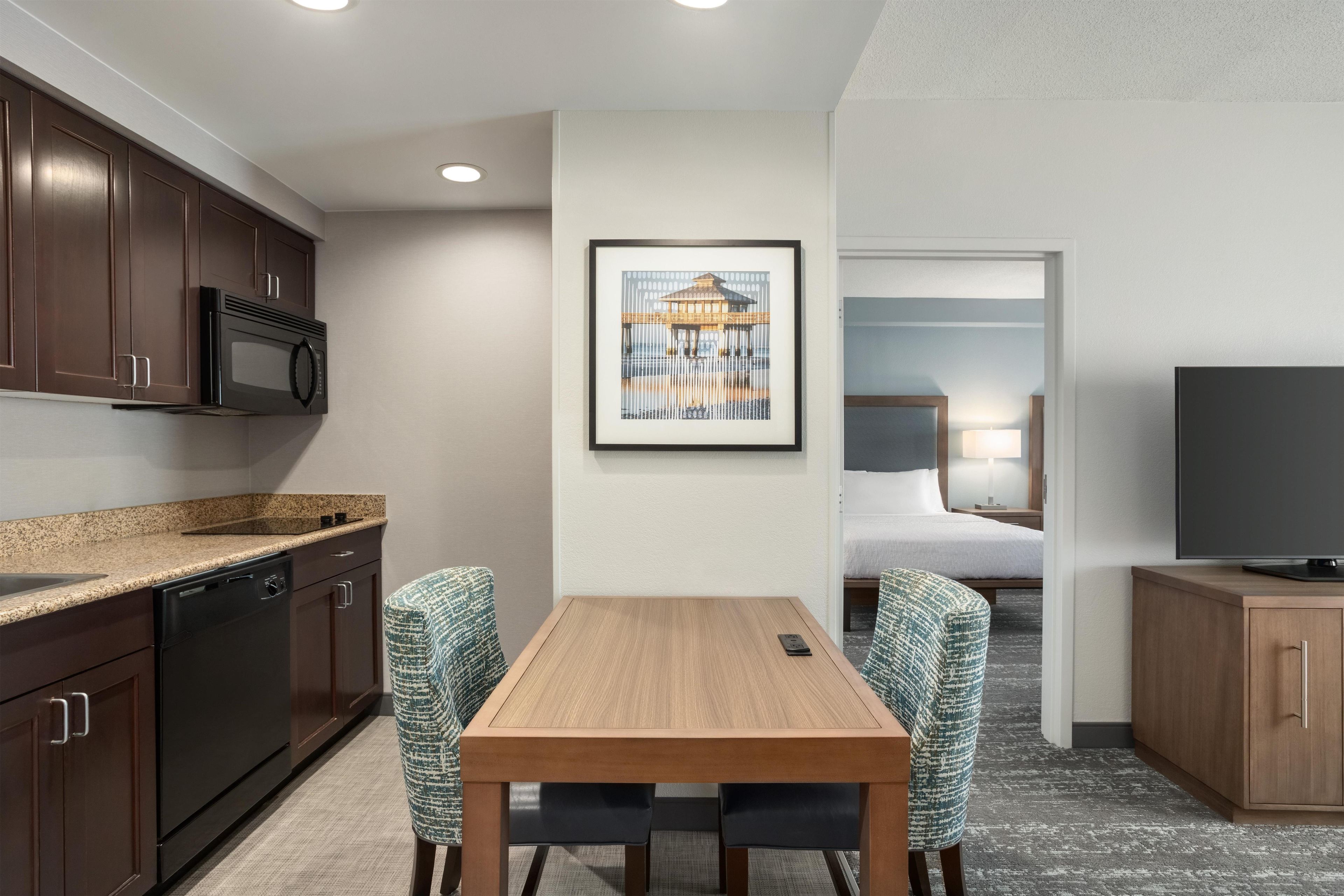 A compact hotel room with a kitchenette, a dining area with two chairs, a bed, a TV, and a framed picture on the wall.