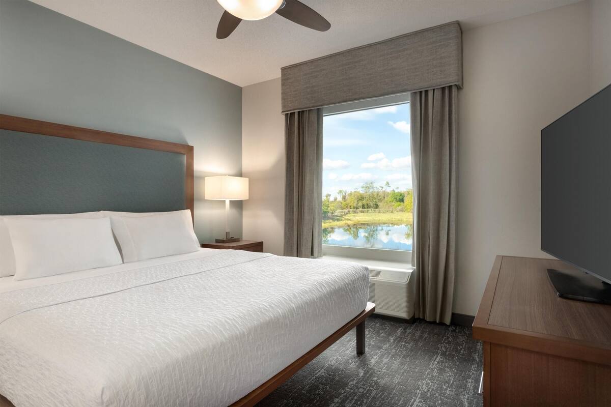 Habitación de hotel con cama, televisor, ventana con vistas a un lago, ventilador de techo y lámpara.
